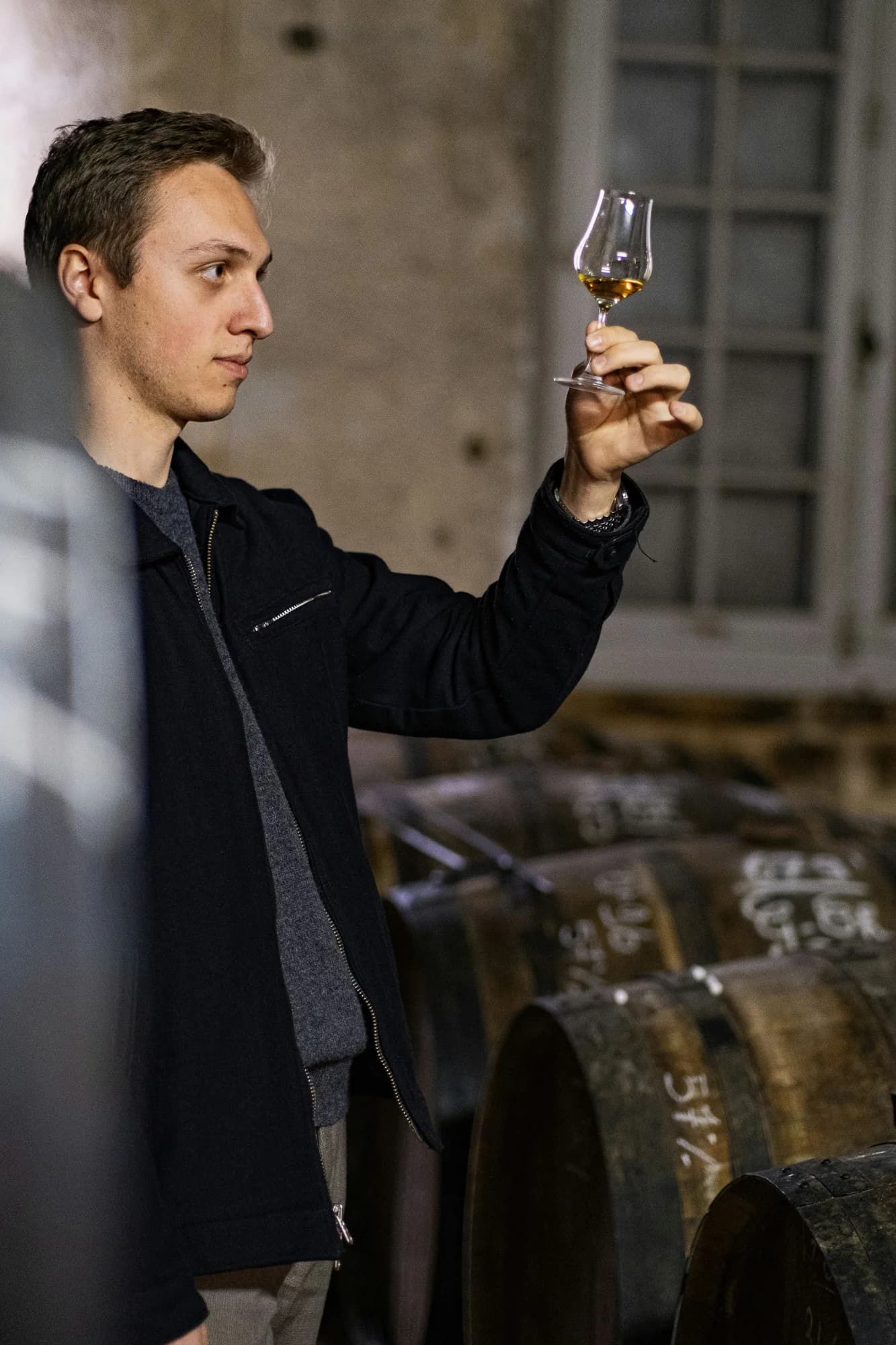 A blender raises a tulip glass of cognac to the light in a cask cellar